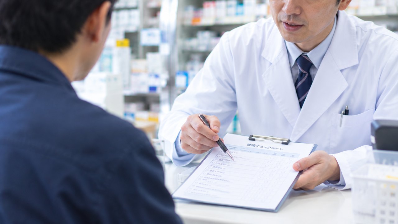 薬局のカウンターで、薬剤師がチェックシートを見せながら男性に説明している様子